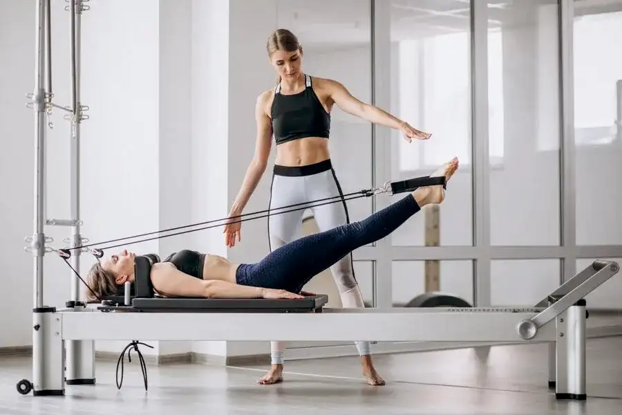 Reformer pilates aleti üzerinde egzersiz yapan kadın ve yanında rehberlik eden pilates eğitmeni. Eskişehir'deki stüdyoda kişisel reformer pilates dersi.